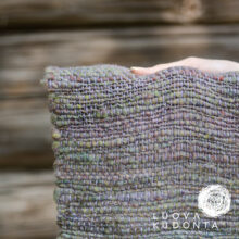 kanervan värinen käsinkudottu huivi kädessä, taustalla näkyy ruskeaa hirsiseinää.