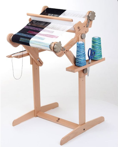 Rigid-heddle-loom-jalustan-hyllyt-luova-kudonta Pirtakangaspuut jalustaan kiinnitettynä, hyllyillä sukkula, sakset ja kaksi lankakartiota.